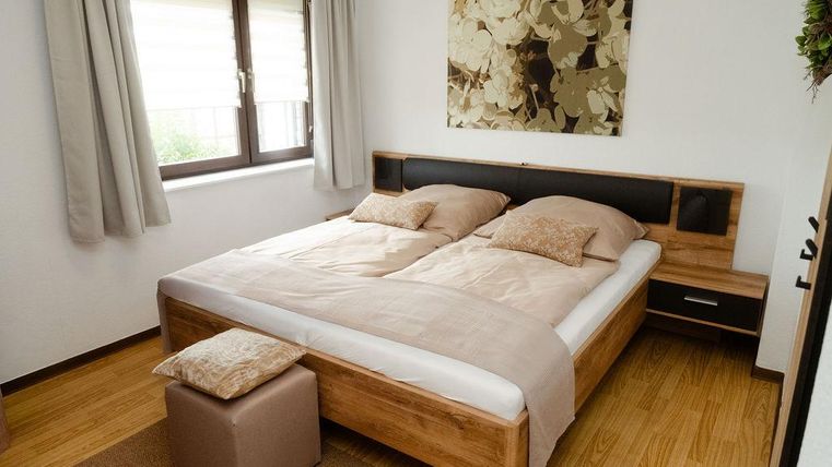 Une chambre moderne avec un grand lit et des coussins beiges. Sur le mur, il y a une œuvre d'art décorative, tandis que des rideaux lumineux éclairent la pièce.