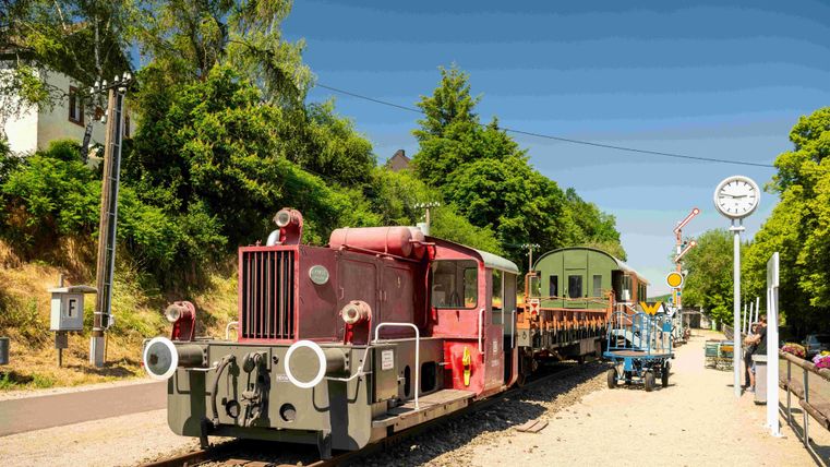 A red locomotive is standing on a railroad track, behind it is a carriage. In the background, there are trees and a station visible.