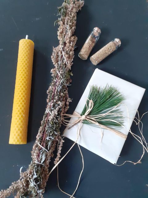 A yellow candle, dried herbs, two small glass vials filled with contents, and a small package decorated with a green twig and string are resting on a dark background. The arrangement appears nature-connected and creative.