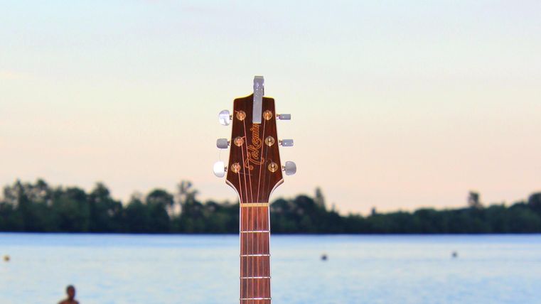 Eine Gitarre steht im Vordergrund vor einem ruhigen Gewässer. Im Hintergrund ist eine Person im Wasser zu sehen, während der Himmel sanft gefärbt ist.