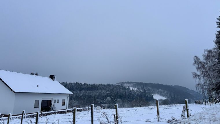 A snow-covered landscape with a white house in the foreground. In the background, there are trees and a cloudy sky.