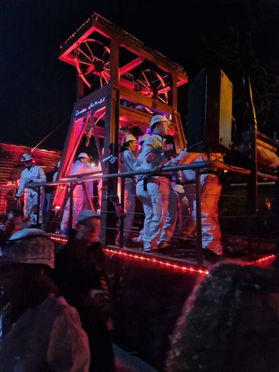 Eine Gruppe von Menschen in weißen Anzügen tanzt auf einer Bühne, die mit roten Lichtern beleuchtet ist. Es ist Nacht und die Atmosphäre ist festlich.