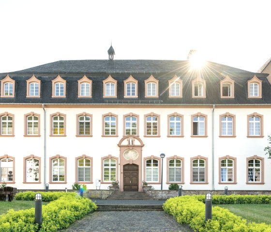 Een historisch gebouw met een symmetrische gevel, veel ramen en een goed onderhouden tuin. De zon schijnt vanachter het dak., © Eifel Tourismus GmbH, Dominik Ketz