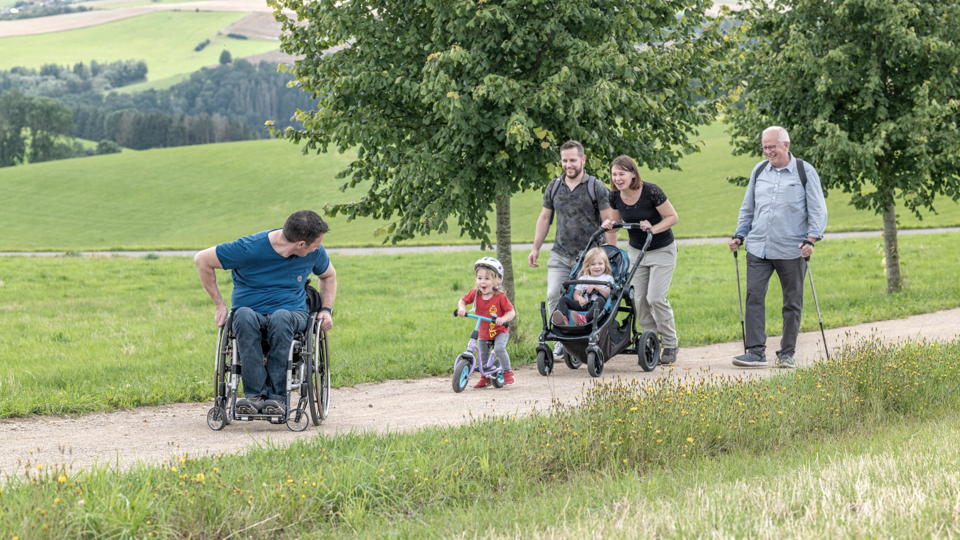 Opa mit Gehstöcken, Eltern, ein Kind mit Laufrad, ein Kind im Kinderwagen und ein Mann im Rollstuhl sind gemeinsam auf dem Komfort-Weg in Ammeldingen bei Neuerburg unterwegs., © Naturpark Südeifel / Thomas Urbany