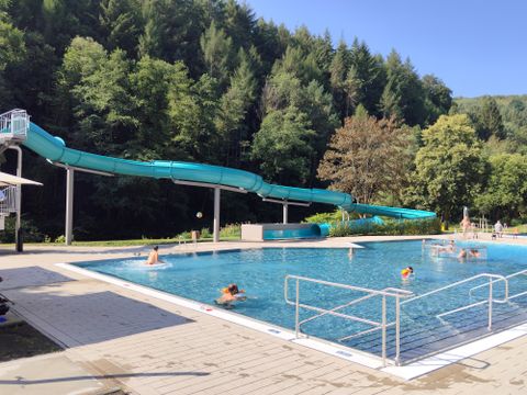 Une piscine avec un long toboggan aquatique et de nombreux arbres en arrière-plan. Quelques personnes nagent et se détendent au bord de la piscine.