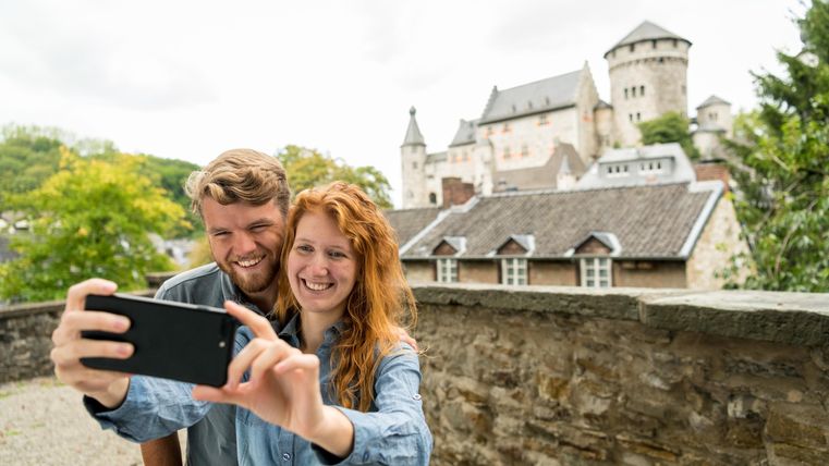 A couple is taking a selfie in front of an old castle. In the background, there are trees and historical buildings.
