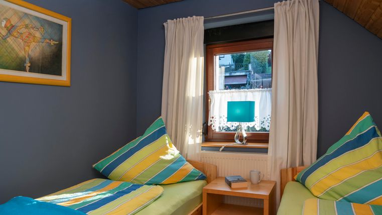 Een gezellige kamer met twee eenpersoonsbedden en kleurrijke, gestreepte dekens. Een raam met gordijnen en een lamp zorgt voor een lichte sfeer.