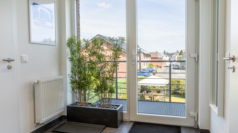 Een heldere entree met grote ramen en uitzicht op de straat. Planten en deurmatten zorgen voor een uitnodigende sfeer.