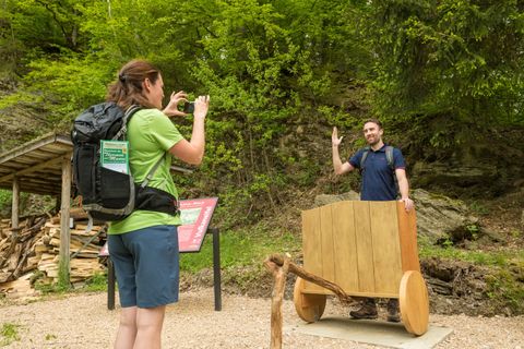 A woman takes a photo of a man standing behind a wooden wagon on a hiking trail in the forest.