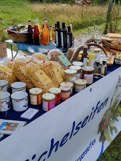 Ein bunter Stand mit verschiedenen regionalen Produkten, darunter Marmeladen, Getränke und Pasta. Im Hintergrund sind Wiesen und Berge sichtbar.