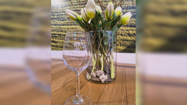 An elegant wine glass stands on a wooden table in front of a vase with fresh, white tulips. In the background, a gentle landscape image can be seen.