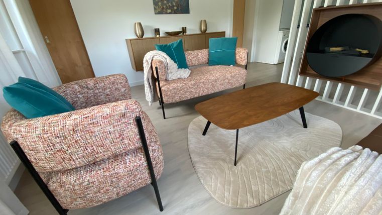 Un salon moderne avec deux fauteuils confortables, une table en bois et une palette de couleurs claires. À l'arrière-plan, des éléments décoratifs et un tapis sont visibles.