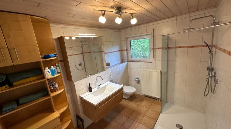 Ein modernes Badezimmer mit hellen Holzmöbeln und einer großen Dusche. Die Wände sind gefliest und es gibt ein Fenster, das Tageslicht hereinlässt.