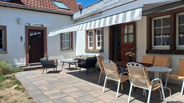 Un espace extérieur confortable avec un sol pavé et plusieurs sièges. Le parasol fournit de l'ombre devant une maison lumineuse avec une porte accueillante.