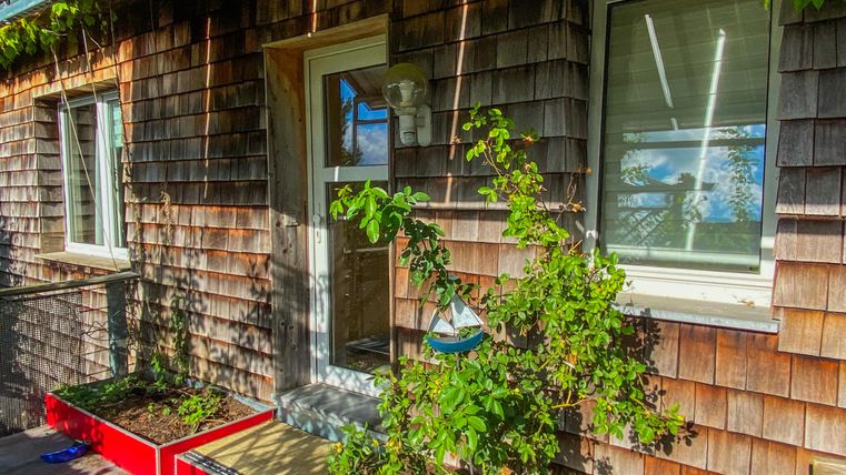 Een met hout bekleed huis met een ingang en twee ramen. Voor het huis staan rode plantenkasten met groene planten.