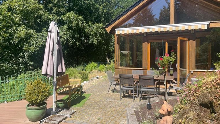 A cozy house with large windows and a terrace. The garden is green with plants and a table with chairs.