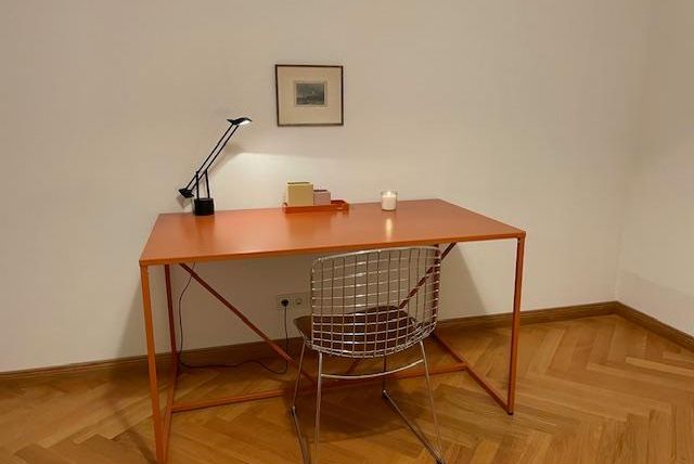 Ein moderner Schreibtisch in leuchtendem Orange mit einer minimalistischen Lampe und einer Drahtstuhl. Im Hintergrund hängt ein kleines Bild an der Wand.