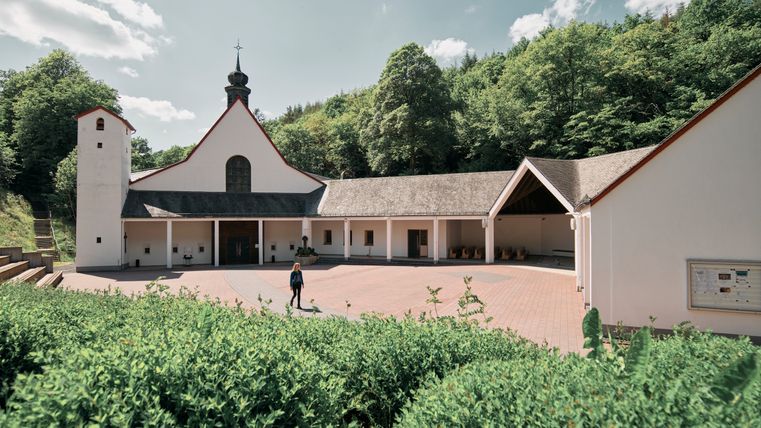 A white-painted church lies on the edge of the forest, surrounded by other buildings and a gently rising, green landscape.