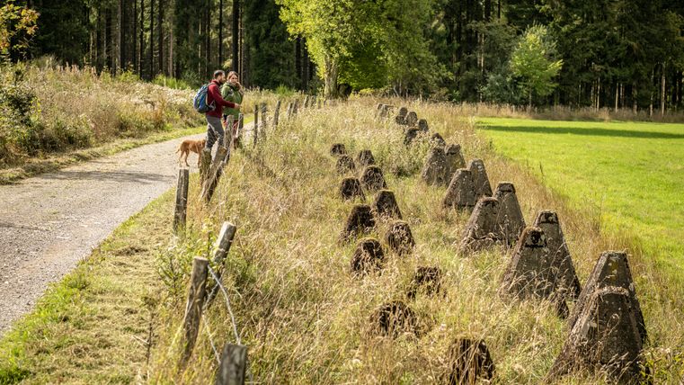 Deux personnes et un chien sur un chemin à côté de barrages antichars dans un environnement rural.