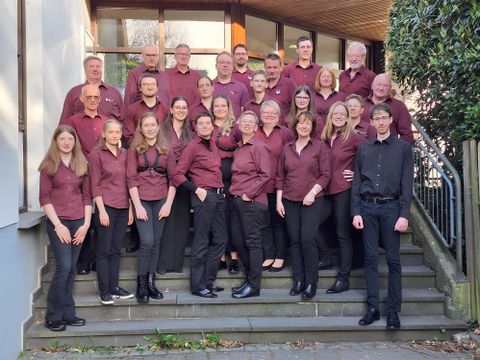 Eine Gruppe von Menschen steht auf einer Treppe vor einem Gebäude. Sie tragen alle einheitliche burgunderfarbene Hemden und schwarze Hosen.