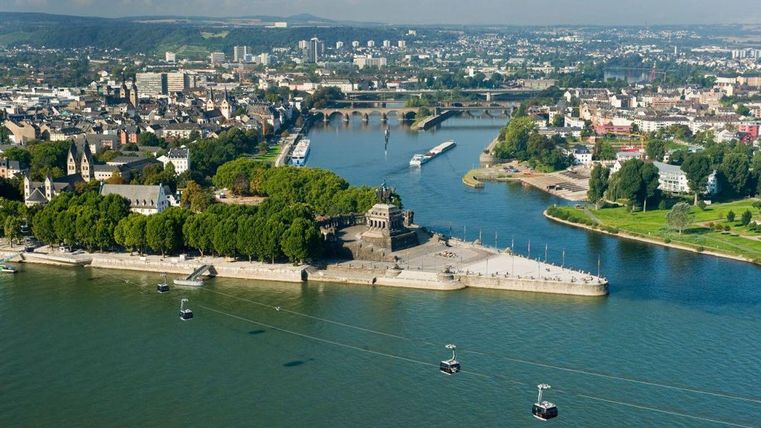 Een luchtfoto van een rivier met een stad op de achtergrond. Op de voorgrond zijn een standbeeld en kabelbanen te zien, omringd door bomen.