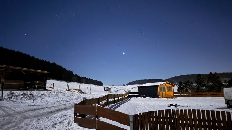 A snowy landscape under a clear night sky. A small wooden house and a fence are visible in the foreground.