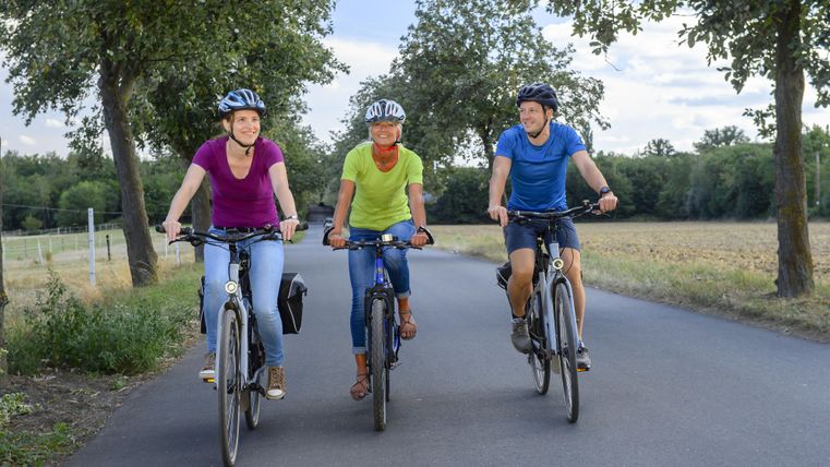 Zwei Radfahrerinnen fahren lächelnd einen Radweg entlang. Im Hintergrund sind grüne Wiesen und Bäume sichtbar.