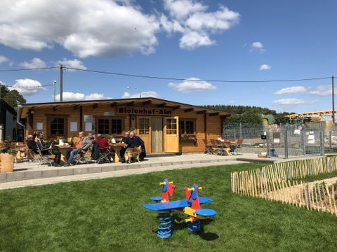 In the background, there is a large single-story wooden hut. In the middle, there is a double door. Above it hangs a wooden sign with the inscription "Bielenhof Alm". On the right and left of the door, there are wing windows with flower boxes. In front of the hut, there is a paved terrace with several tables and chairs. Two of the tables are occupied by a group of men. In the foreground, there is a spring rider on a grassy area in the colors blue, yellow, and red.