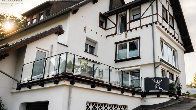 Ein modernes Haus mit einem großen Balkon und Glasgeländer. Die Fassade ist hell gestrichen und zeigt traditionelle Dachdetails.