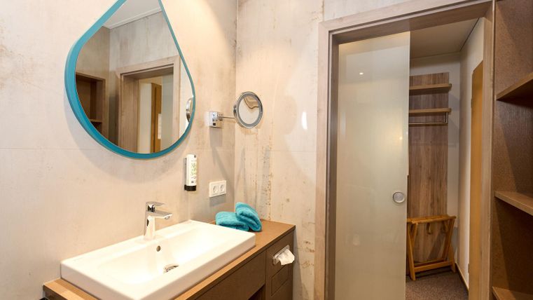 Een moderne badkamer met een witte wastafel en een grote, ronde spiegel. De ruimte is licht en minimaal ingericht, met een houten plank en een douchecabine.