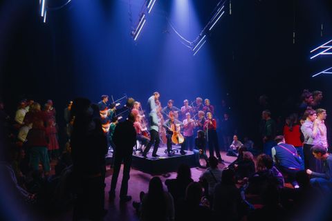 A lively event with musicians on a stage and an engaged crowd. Colorful lights and a cheerful atmosphere create an inviting vibe.