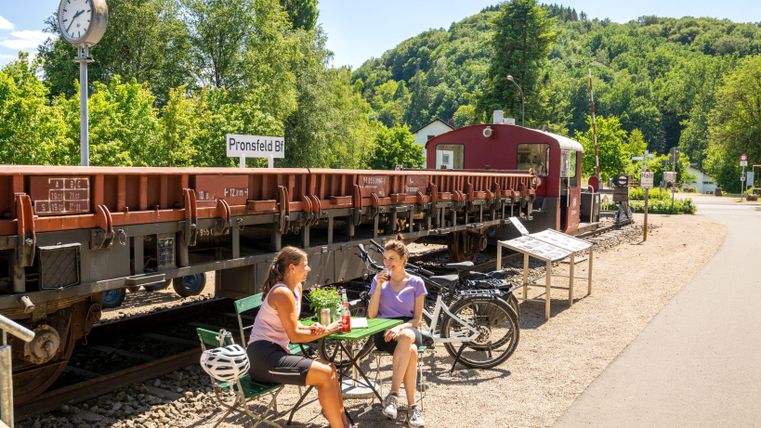 Zwei Personen sitzen an einem Tisch in der Nähe eines Bahnhofs. Hinter ihnen steht ein Waggon und die Umgebung ist von grünen Hügeln umgeben.