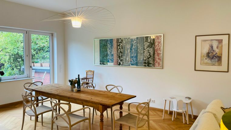 Une salle à manger lumineuse avec une longue table en bois et plusieurs chaises. Au mur, quelques œuvres d'art colorées sont accrochées, et de grandes fenêtres laissent entrer beaucoup de lumière naturelle.