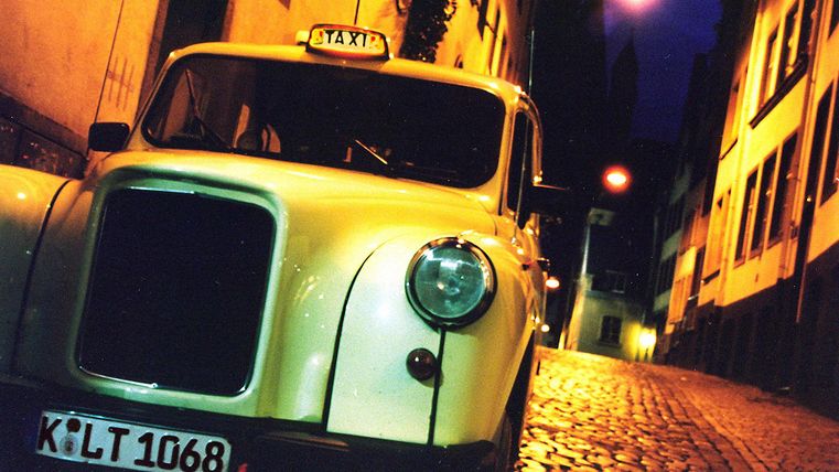 A classic taxi is standing in a narrow, cobblestone street. The street lamps create an atmospheric evening ambiance.
