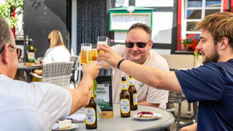 Een vrolijke groep vrienden die proost op een biertje. Op de achtergrond zijn stoelen en tafels te zien in een uitnodigende biergarten.