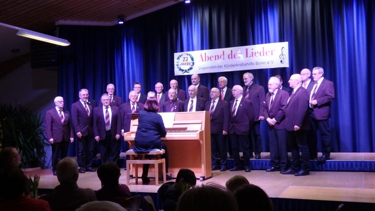 A men's choir performs on a stage while a pianist accompanies them. In the background, there is a banner reading "Evening of Songs."