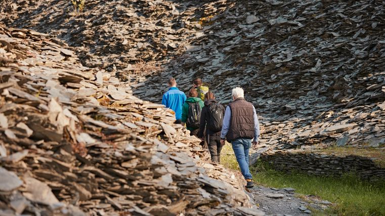 Un groupe de cinq personnes marche le long d'un sentier étroit, entouré de pentes rocailleuses. Le paysage est verdoyant et invite à la découverte.