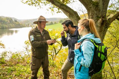 Ein Ranger mit einem Hut erklärt zwei Besuchern die Natur im Nationalpark Eifel