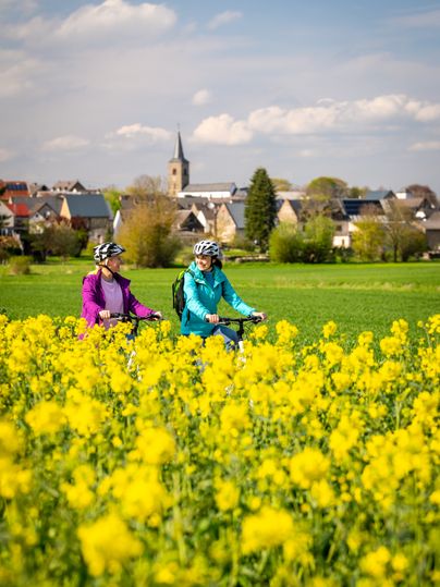 Zwei Radfahrerinnen fahren durch ein blühendes Rapsfeld mit einem Dorf im Hintergrund.