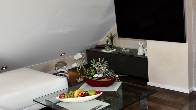 A modern living room with a large television and a glass table. On the table, there is a fruit bowl and some plants.