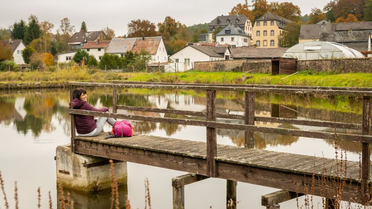 Persoon zittend op een steiger aan het water met een rugzak, gebouwen en bomen op de achtergrond.