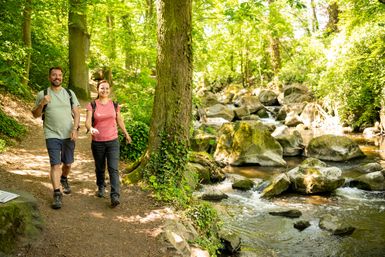 Zwei Personen wandern auf einem schmalen Pfad. Neben dem Pfad fließt ein Fluss.