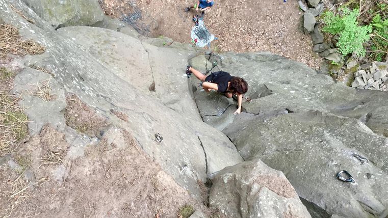 Een persoon klimt aan een steile rotswand. Op de achtergrond zijn andere personen en een bebost gebied zichtbaar.