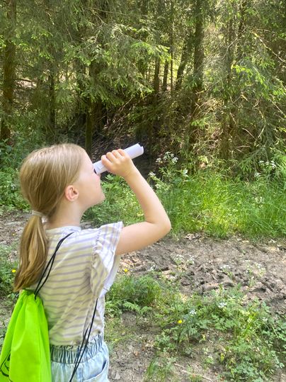 Ein Kind mit einem grünen Rucksack schaut durch ein selbstgemachtes Fernglas in einem Wald.