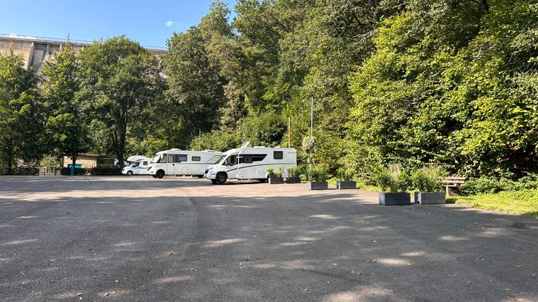 Een rustige parkeerplaats met campers, omringd door groene bomen. De lucht is helder en blauw.