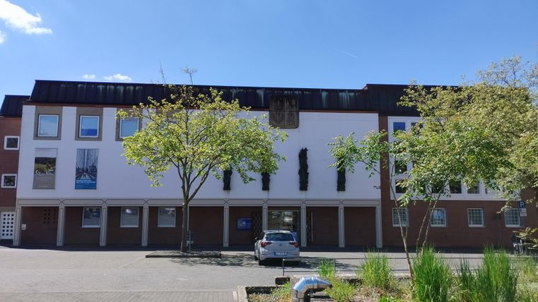 Un bâtiment moderne avec de grandes fenêtres et un ciel bleu clair. Au premier plan, on peut voir des arbres et un stationnement.