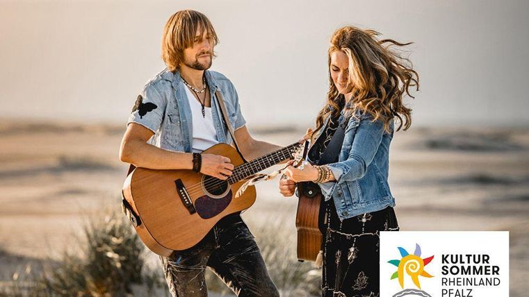 Ein Paar spielt Musik am Strand mit Gitarren. Der Hintergrund zeigt Sanddünen und einen klaren Himmel.