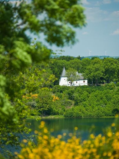 Ein weißes Gebäude mit Türmen liegt malerisch an einem See, umgeben von grüner Natur. Die Szenerie strahlt Ruhe und Idylle aus.