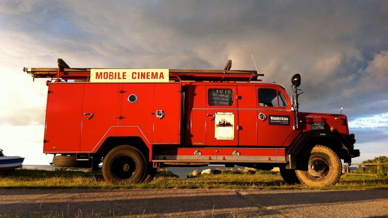 A red vehicle with the inscription "Mobile Cinema." It is standing on a street under a cloudy sky.