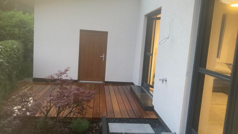 Eine moderne Hausansicht mit einem kleinen Holzdeck und einer Eingangstür. Im Vordergrund sind einige Pflanzen und der Weg aus Platten zu sehen.